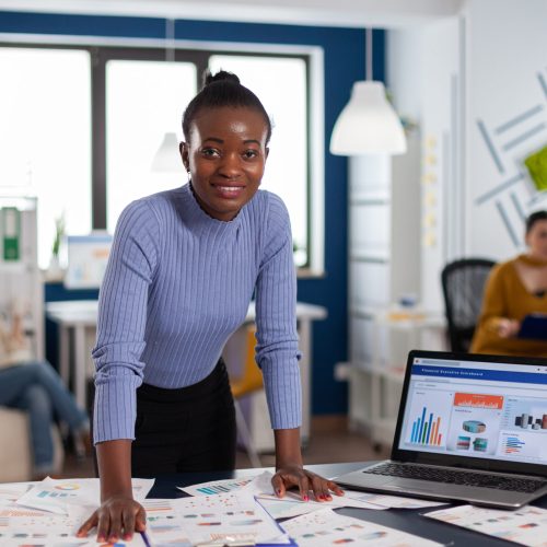 Dark skinned woman and colleagues in corporate start up office working to finish project. Diverse team of business people analyzing company financial reports from computer.