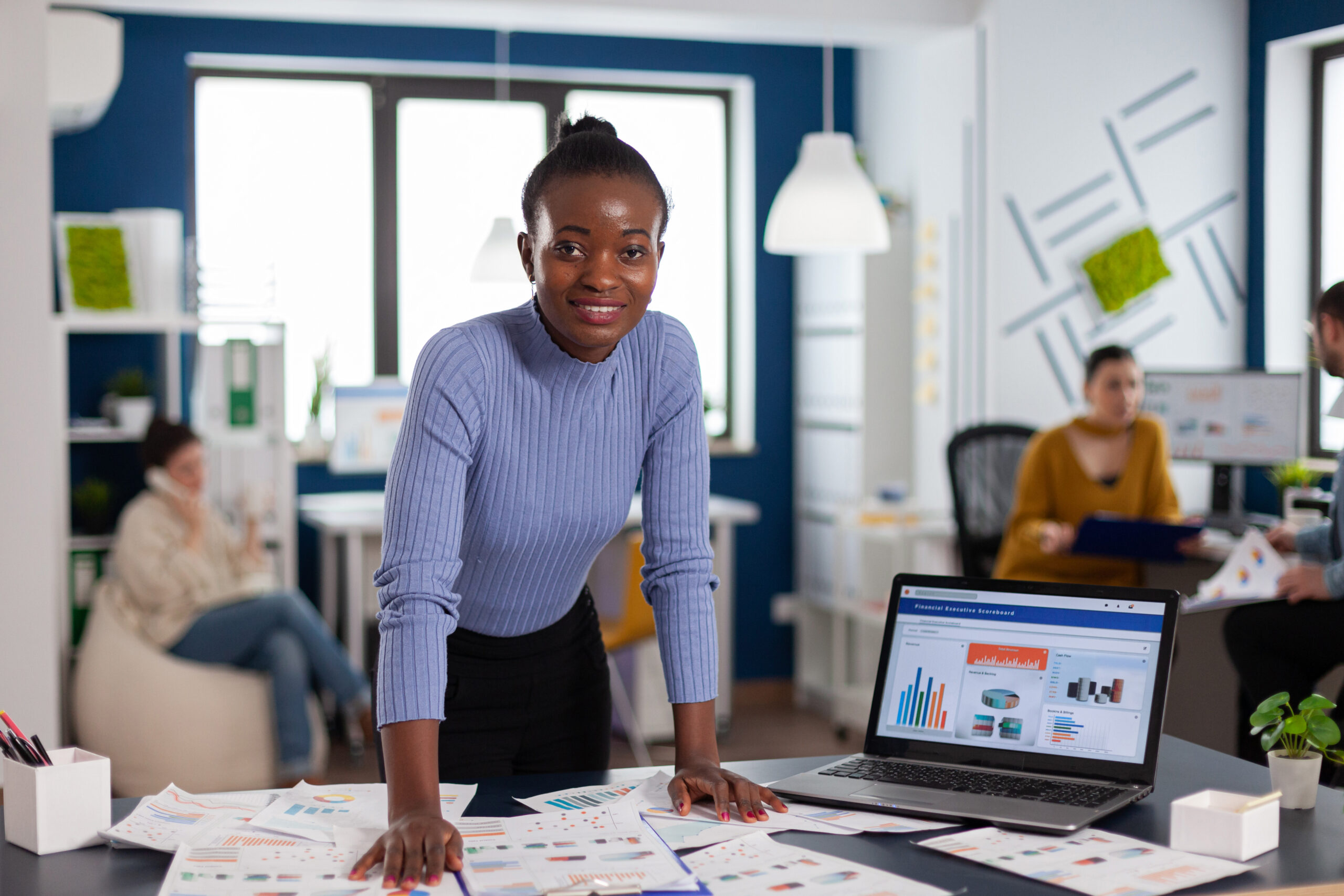 Dark skinned woman and colleagues in corporate start up office working to finish project. Diverse team of business people analyzing company financial reports from computer.